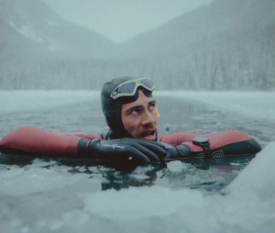 Connor Emeny in a neoprene hood and goggles, resting his arms on the ice at the edge of a frozen mountain lake during a cold-water swim