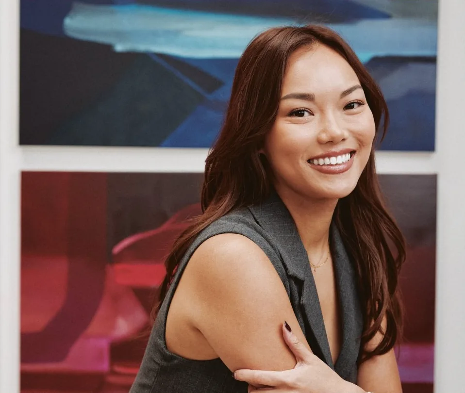Annika Ng, co-founder and COO of Blossom Social, smiling in a grey sleeveless blazer in front of large blue and red abstract paintings