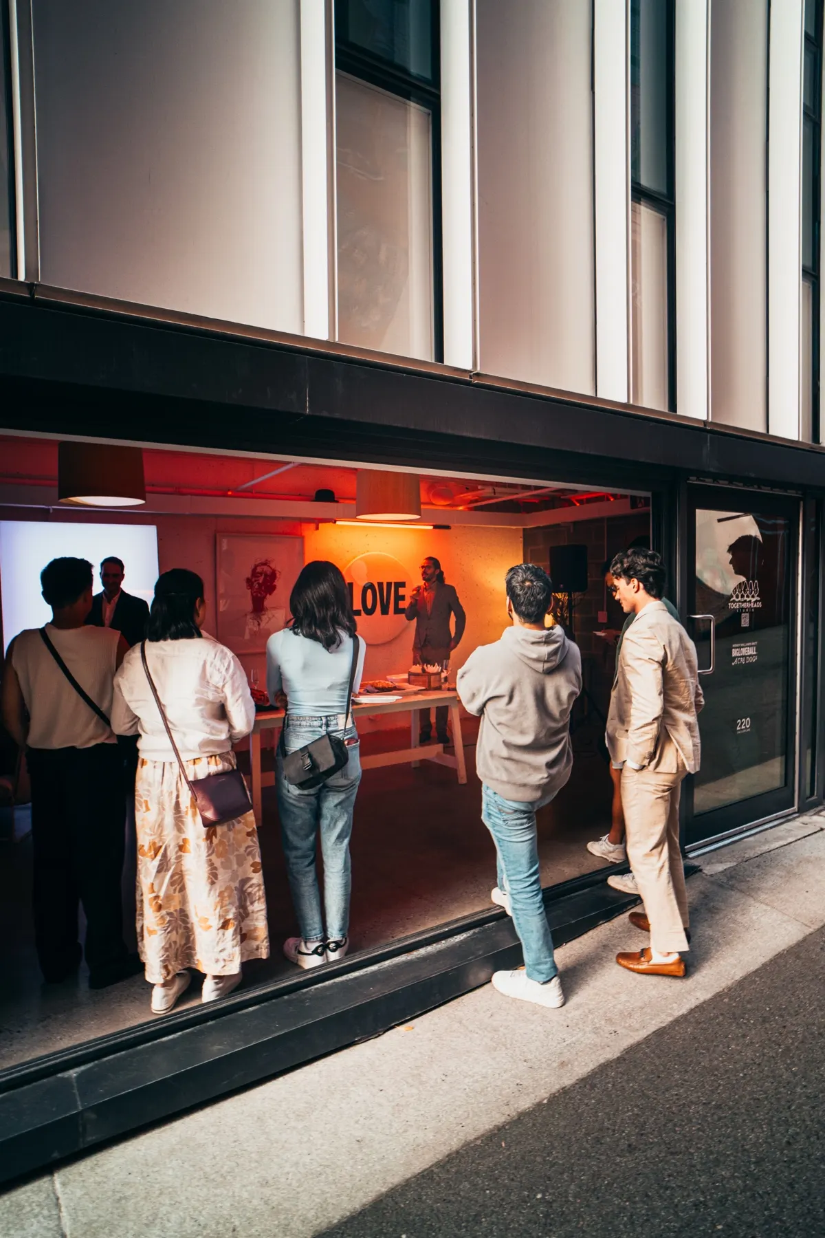 People on the street looking in through the open glass at a Founder Feast evening on July 30, 2026