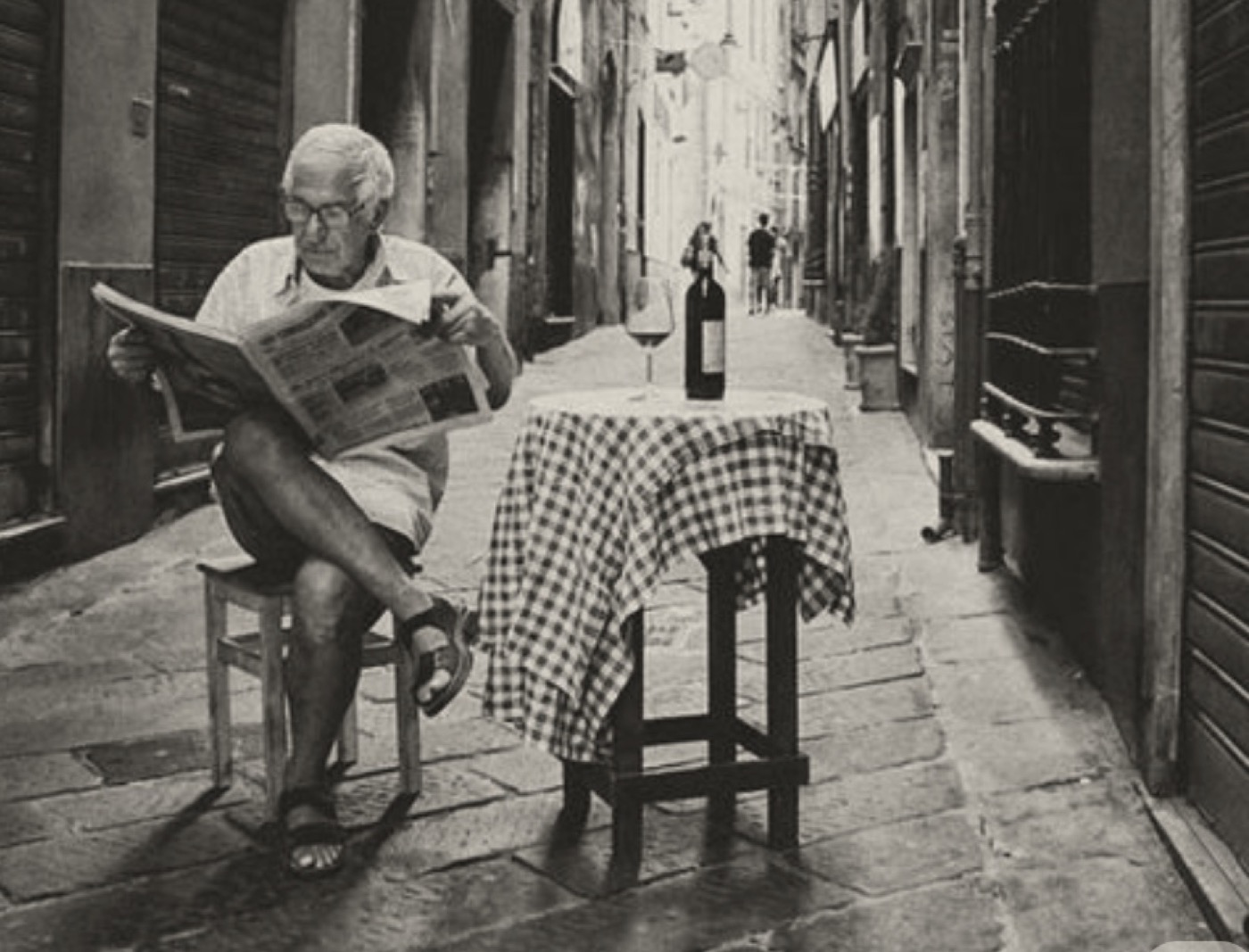 A man reading a newspaper at a small table with a checked cloth in a narrow stone street.