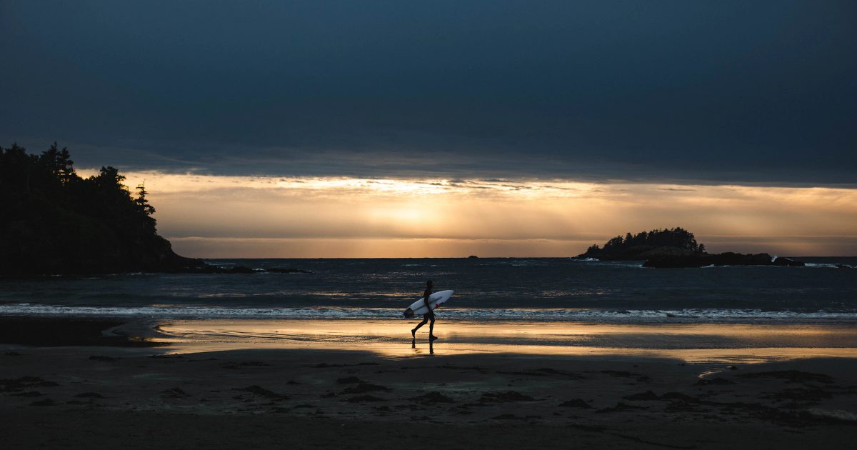 A surfer walking a West Coast beach at sunset.